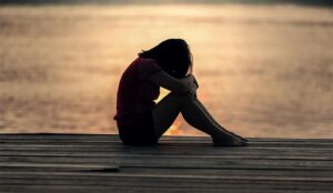 Flicka som sitter hopkrupen i sorg på en brygga vid vatten. Foto: Pexels-Pixabay