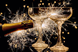 Två glas som skålar och en champagneflaska som sprutar ut sitt innehåll. Guldglittrigt mot svart bakgrund. Foto: Myriams Fotos, Pixabay