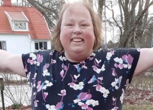 Marina Sängängel, ljushårig kvinna, halvporträtt, blåblommig blus. I bakgrunden hus med rött tegeltak..