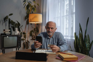 En ensam man som sitter i sitt hem och tittar i sin mobil. Gammal TV i i rummet. En grönskande växt.