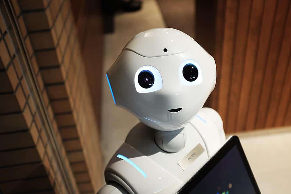 En vit robot, ansikte och del av överkroppen. Man ser en del av en datorskärm som den bär på. Det står i ett rum där bakgrunden består av bruna tegelväggar.