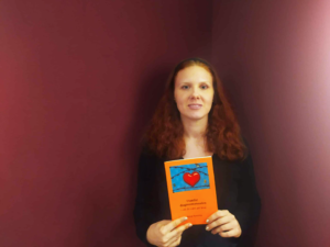 Anna Nyström håller i sin bok som är orange. Bakgrunden är vinröd.