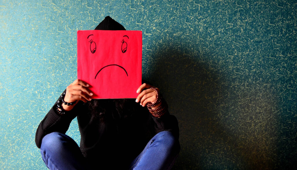 Pojke som sitter med benen i kors mot en grön bakgrund. Framför sig har han ett rött paper med en smiley som visar en ledsen gubbe. Foto: PDPics, Pixabay