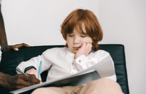 Pojke med vit skjorta som sitter med skrivbok. En hjälpande hand med penna från vänster. Foto: Mikhail Nilov från Pexels