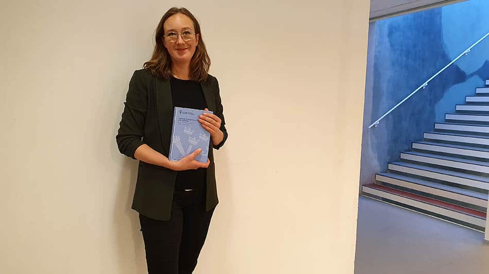 Forskaren Cecilia Åkesdotter på GIH med sin avhandling. Hon är mörkklädd och står mot en beige vägg. En trappa syns till höger.