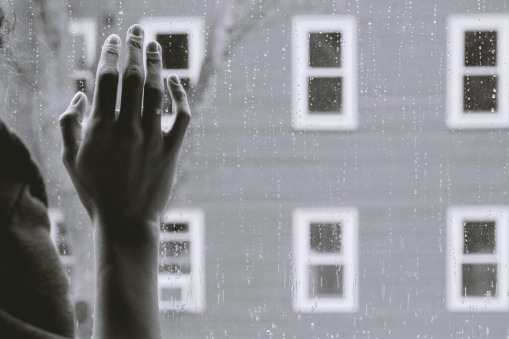 Kvinna som håller hand mot fönster, husfasad i bakgrunden. Svartvit bild.